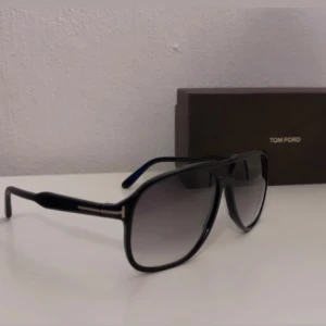 Svarta Tom Ford pilotglasögon - Snygga svarta pilotglasögon från Tom Ford med klassisk design och mörka tonade linser. Ramen är i svart syntetmaterial med diskreta gulddetaljer vid skalmarna. Perfekt för dig som vill ha en stilren och exklusiv look. Äkthetsbevis finns! 