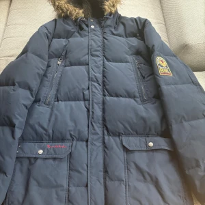 Mörkblå dunjacka Bondelid med päls - Snygg mörkblå dunjacka från Bondelid med huva och fuskpäls. Jackan har två dragkedjefickor på bröstet, två fickor med knapp, patch på ärmen och quiltad design. Insidan av huvan är fodrad med mjuk teddy. Perfekt för kalla dagar.