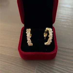 Glittriga guldiga creoler - Säljer ett par snygga creolörhängen i guldton med glittriga stenar runt hela ringen. Örhängena har en modern design med rektangulära stenar som ger extra bling. Perfekta för dig som vill sticka ut och addera lite lyx till din look. Guldpläterade i 14k, röda boxen ingår INTE!