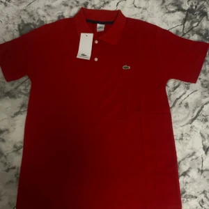 Röd pikétröja från Lacoste - Snygg röd pikétröja från Lacoste med klassisk krage och två vita knappar framtill. Tröjan har det ikoniska gröna krokodilmärket broderat på bröstet. Tillverkad i mjuk bomull som känns skön mot huden. Perfekt för dig som gillar stilren och sportig look.