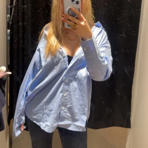 Oversized ljusblå skjorta - Säljer en oversized ljusblå skjorta med klassisk krage och knäppning framtill. Skjortan har långa ärmar och en avslappnad passform, perfekt att styla lager på lager eller bära öppen över ett linne. Köpte i storlek XS men är väldigt oversized som man tydligt ser. Endast använd en gång!!