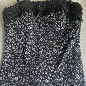 Svart glittrig klänning med volanger - Supersnygg svart klänning med smala axelband, glittrigt leopardmönster och spetsdetaljer upptill. Kjolen har flera lager volanger i transparent tyg som ger en cool och lekfull look. Perfekt för dig som vill sticka ut med en unik och trendig stil.passar storlek 38-40