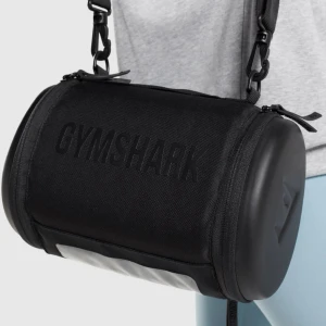 Svart axelväska från Gymshark - Cool svart axelväska från Gymshark i cylinderformad design. Väskan har tydlig logga, justerbar axelrem och dragkedja upptill. Tillverkad i slitstark polyester med detaljer i syntet och hårda gavlar. Perfekt för att bära med sig essentials till gymmet eller på språng. Helt ny och aldrig använt. 