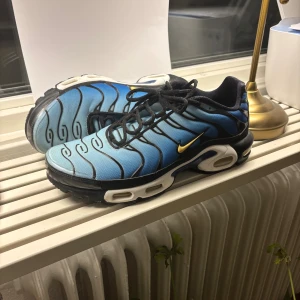 Nike Air Max Plus blå och svart - Nike Air Max Plus sneakers i blått och svart med coola vågformade linjer och gul swoosh på sidan. Skorna har en robust platt sula med synliga luftkuddar och svarta snören. Ovandelen är i syntetmaterial och mesh för bra ventilation. Perfekt för dig som gillar streetwear och sportig stil.