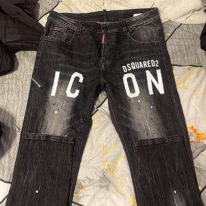 Svarta Dsquared2 ICON jeans - Säljer ett par svarta Dsquared2 jeans med vit 'ICON'-print och Dsquared2-logga framtill. Jeansen har en tvättad look med vita färgstänk och klassisk femficksdesign. Materialet är jeans och passformen är normal. Perfekta för dig som vill sticka ut med streetstyle.
