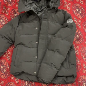 Svart pufferjacka från Canada Goose - Svart pufferjacka från Canada Goose med huva och klassisk logga på ärmen. Jackan har dragkedja och tryckknappar framtill, två stora fickor och quiltad design. Perfekt för kalla dagar med sin tjocka och fluffiga look.