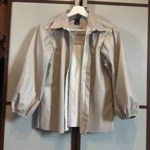 Beige overshirt från Lindex XS - Snygg beige overshirt från Lindex i storlek XS. Skjortan har bred krage, ballongärmar med resår vid ärmslut och knappar framtill. Materialet är syntet med en lätt glansig finish som ger en cool look. Perfekt att styla lager på lager.