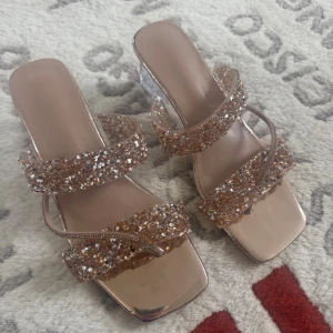 Glittriga sandaler med transparent klack - Säljer ett par sandaler med fyrkantig tå och transparent klack. Remmarna är täckta av glittriga paljetter i guld och silvertoner som ger extra bling. Snyggt metallicbeige material på sulan och ovandelen. Perfekt för dig som vill sticka ut med lite extra glam.