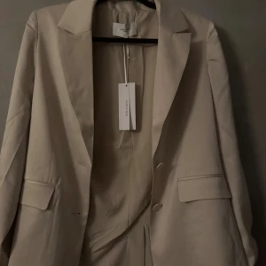 Satin kavaj  - Stilren beige kavaj från House of Dagmar med klassisk krage, två framfickor med lock och två knappar framtill. Kavajen har en rak passform och är tillverkad i ett slätt material som ger en elegant look. Perfekt för dig som gillar minimalistisk stil.