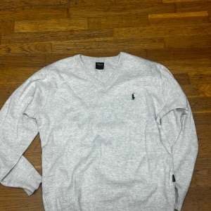  Polo Ralph Lauren tröja - Ljusgrå v-ringad tröja från Polo Ralph Lauren i storlek L. Klassisk design med broderad logga på bröstet och ribbade muddar vid ärmslut och nederkant. Perfekt för dig som gillar stilrena och tidlösa plagg. Materialet känns mjukt och skönt mot huden.