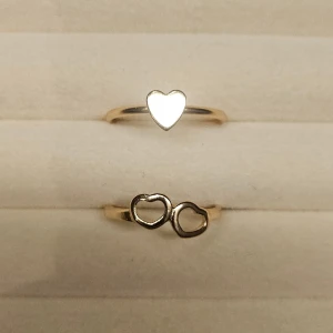Två guldiga ringar med hjärtan - Säljer två tunna guldiga ringar, en med ett fyllt hjärta och en med två öppna hjärtan. Perfekta att bära tillsammans eller var för sig för en söt och romantisk look.