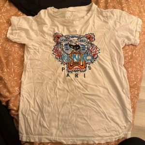 Vit Kenzo t-shirt med tigertryck - Vit t-shirt från Kenzo med ikoniskt broderat tigerhuvud i blått, orange och turkost framtill. Klassisk passform och rund halsringning. Perfekt statement-plagg för dig som gillar streetstyle och coola prints.