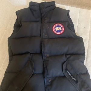 Svart dunväst från Canada Goose XS - Svart dunväst från Canada Goose med klassisk logga på bröstet och broderad etikett på fickan. Västen har hög krage, två snedställda fickor och dragkedja framtill. Perfekt för kalla dagar när du vill ha både stil och värme.