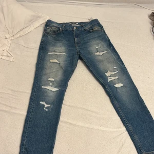 Blå ripped jeans från Zara, strl 44 - Säljer ett par blå jeans från Zara i storlek 44 med snygga slitningar och ripped detaljer framtill. Modellen har klassisk femficksdesign, normal passform och raka ben. Jeansen är tillverkade i bomull och har en cool, avslappnad vibe.