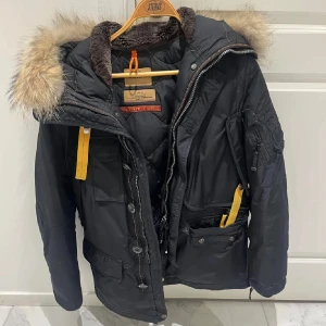 Parajumpers svart parkajacka XS - Svart parkajacka från Parajumpers med päls på huvan, flera fickor och ikonisk gul rem vid kragen. Jackan har dragkedja, knappar och detaljer i orange. Perfekt för kalla vinterdagar och har en snygg, sportig look.