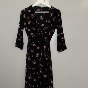Svart blommig omlottklänning Forever 21 - Svart omlottklänning från Forever 21 med rosa och vita blommor. Klänningen har trekvartsärmar, v-ringning med volangdetalj och knyt i midjan. Lätt och flowy material som ger en snygg siluett. Perfekt för dig som gillar romantiska vibes och blommigt.