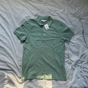 Grön pikétröja från Lacoste slim fit - Snygg grön pikétröja från Lacoste i slim fit-modell. Klassisk krage, korta ärmar och knäppning framtill med tre knappar. Ikoniska krokodilloggan broderad på bröstet. Tillverkad i mjuk bomull som känns skön mot huden.