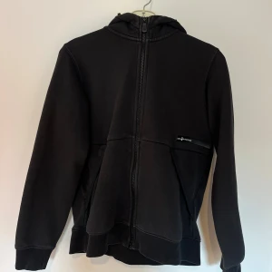 Svart hoodie från Sail Racing - Svart hoodie från Sail Racing med hel dragkedja framtill, huva och två fickor. Tröjan har en stilren look med diskret logga på bröstet och är tillverkad i mjukt bomullsmaterial. Perfekt för chill dagar eller när du vill ha en enkel och snygg stil.