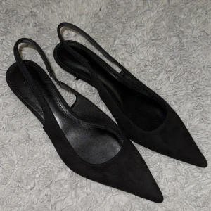 Svarta pumps med spetsig tå från H&M - Snygga svarta pumps från H&M i storlek 38 med spetsig tå och öppen häl. Skorna har en elegant och stilren design med smal rem runt hälen. Materialet är mocka-liknande och sulan är i syntet. Perfekta för dig som gillar minimalistisk och trendig stil.
