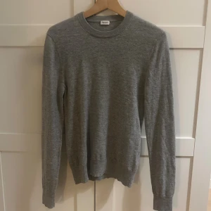 Grå stickad tröja från Filippa K - En stilren grå stickad tröja från Filippa K med rund halsringning och långa ärmar. Tröjan är i mjuk ull och har ribbade muddar vid ärmslut och nederkant. Perfekt för lager-på-lager under kyliga dagar.