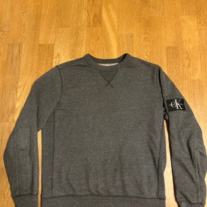 Calvin Klein sweatshirt  - Mörkgrå sweatshirt från Calvin Klein Jeans med klassisk rund hals och ribbade muddar. Stor CK-logga på vänster ärm ger en snygg streetkänsla. Perfekt för dig som gillar stilrena och bekväma plagg.