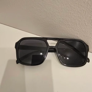 Svarta solglasögon med bred båge - Snygga svarta solglasögon med bred, fyrkantig båge och mörka glas. De har en modern och stilren design med en cool detalj över näsryggen. Perfekta för att ge din outfit en extra edge.