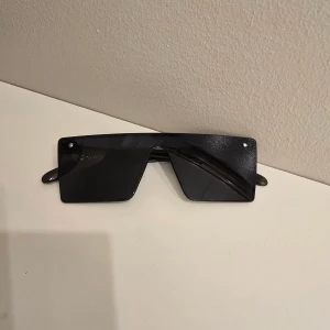 Svarta futuristiska solglasögon - Säljer ett par svarta solglasögon med rektangulär, futuristisk design och breda bågar. Glasögonen har en helsvart look och ger en riktigt edgy vibe, perfekta för dig som vill sticka ut i mängden.