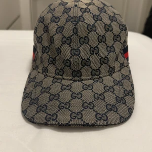Gucci keps med GG-mönster och röd-blå rand - Snygg keps från Gucci med klassiskt GG-mönster i mörkblått på beige bakgrund. Kepsen har en böjd skärm och justerbar rem baktill. På sidan finns en ikonisk röd-blå rand som ger extra lyxig känsla. Perfekt för dig som vill sticka ut med designerstil.
