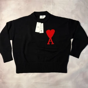Svart stickad tröja från Ami - Svart stickad tröja från Ami Alexandre Mattiussi med röd hjärtapplikation och bokstaven A på bröstet. Tröjan har rund hals, ribbade muddar och en loose passform. Perfekt för dig som gillar stilrena detaljer med en twist.
