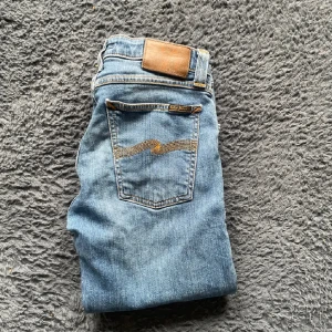Nudie jeans  - Säljer ett par ljusblå skinny jeans från Nudie Jeans med klassiska orange sömmar och femficksdesign. Jeansen har normal passning i midjan och är tillverkade i denim med dragkedja. Perfekta för dig som gillar en smal siluett. Längd 107cm