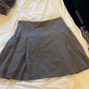 Grå vid kortkjol med veck - Säljer en snygg grå kortkjol med utsvängd passform och markerade veck. Kjolen har en stilren look och är perfekt för dig som gillar minimalistiska plagg. Passar till många olika stilar och är lätt att matcha med både sneakers och boots.