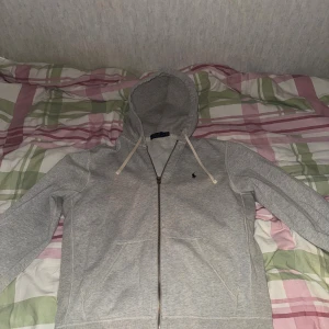 Grå hoodie från Polo Ralph Lauren - Grå hoodie från Polo Ralph Lauren med dragkedja och huva med vita snören. Klassisk logga broderad i mörkblått på bröstet. Tröjan har två fickor framtill och är tillverkad i mjuk bomullsmix. Perfekt för chill dagar och enkel att matcha med jeans.