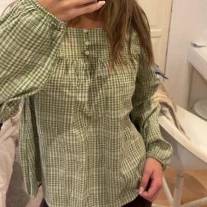 Grön rutig henley från Springfield - Grön och vit rutig henley-skjorta från Springfield med tre knappar framtill. Skjortan har långärmade, lite puffiga ärmar och är gjord i mjuk bomull. Den har en avslappnad passform och fyrkantig halsringning.