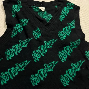 Svart stickad väst med grön text - Cool svart stickad väst med grönt grafiskt textmönster över hela plagget. Västen har v-ringning och ribbade kanter. Perfekt för att ge outfiten en edgy vibe.