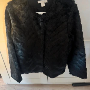 Svart pälsjacka från H&M storlek s  - Säljer en svart pälsjacka från H&M med rund hals och lång ärm. Jackan har en mjuk och fluffig yta i syntetpäls och är perfekt för dig som vill ha en cool och trendig look. Enkel design utan synliga knappar eller dragkedja.
