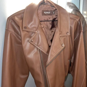 Brun skinnjacka från Bianca x Nelly - Snygg brun skinnjacka från Bianca x Nelly i storlek 36. Jackan har klassisk bikerstil med bred krage, synliga sömmar och silverfärgad dragkedja. Materialet är mjukt och har en matt finish. Perfekt för dig som vill ha en cool och trendig look.