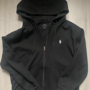 Svart hoodie från Polo Ralph Lauren - Svart hoodie från Polo Ralph Lauren med dragkedja och klassisk vit logga på bröstet. Tröjan har huva med snörning och känguruficka framtill. Perfekt för chill dagar och enkel att matcha med allt. Materialet är mjuk bomull för extra komfort.