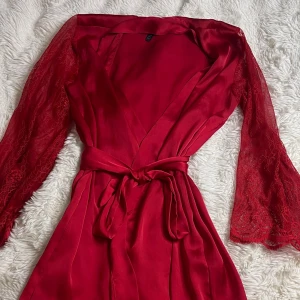 Röd satin morgonrock med spetsärmar  - Röd omlott morgonrock i satinliknande material med långa spetsärmar och knytband i midjan. Klänningen har v-ringning och transparenta ärmar med fina spetsdetaljer. Den är från hunkemöller. Det finns lite spår av brun utan sol på den men är ganska säker det går att få bort på något vis. Nypris :500 kr