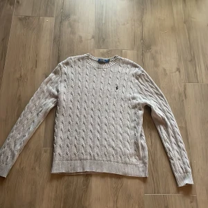 Grå kabelstickad tröja Polo Ralph Lauren - Kabelstickad grå tröja från Polo Ralph Lauren med klassisk rund hals och broderad logga på bröstet. Tröjan har lång ärm och ribbade muddar för en snygg passform. Perfekt för dig som gillar stilren och tidlös design.