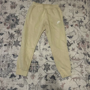 Beige mjukisbyxor från Nike - Beiga mjukisbyxor från Nike med broderad logga på vänster lår. Byxorna har resår och snörning i midjan samt muddar vid bensluten. Tillverkade i mjukt bomullsmaterial för extra komfort. Perfekta för chill eller träning.