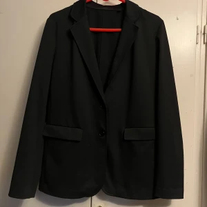 Svart kavaj från Uniqlo - Stilren svart kavaj från Uniqlo med klassisk krage och två knappar framtill. Kavajen har raka ärmar, två fickor och är tillverkad i en mix av polyester, lyocell och elastan för en bekväm passform. Köpte den för några år sedan men har bara använt några gånger.