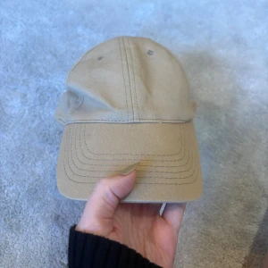 Beige keps med justerbar rem - Snygg beige keps med klassisk böjd skärm och ventilerande hål upptill. Mjuk, bekväm och enkel att justera med kardborreband på baksidan. Stilren design som funkar till alla outfits! Några sminkfläckar på insidan vid pannan som inte syns när man har på sig den. 