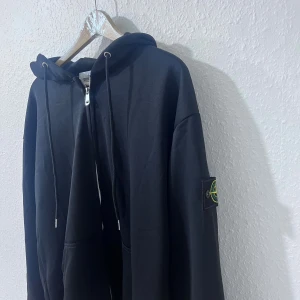 Svart hoodie från Stone Island - Svart hoodie från Stone Island med huva och snörning, dragkedja framtill och klassisk logopatch på ärmen. Tröjan har en loose passform och är tillverkad i mjuk bomull. Perfekt för chill dagar och streetwear-stil.