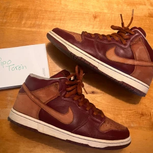 Nike Dunk SB mid 2007 - Säljer ett par Nike Dunk High sneakers i vinrött och brunt läder och mocka. Skorna har klassisk snörning, vit mellansula och mörk yttersula. EU 44 ingen og box och heeldrag som jag lagat