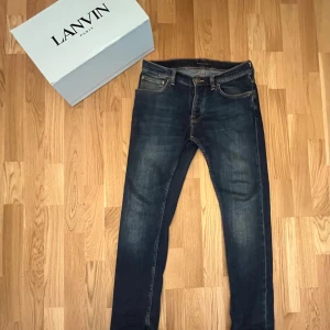 Mörkblå jeans från Nudie Jeans Co - Säljer ett par mörkblå jeans från Nudie Jeans Co med klassisk femficksdesign och snygga slitningar på bakfickorna. Jeansen har orangea sömmar och Nudie-logga på läderpatchen bak. Perfekt passform och stilren look för dig som gillar denim. Modellen är grim Tim 