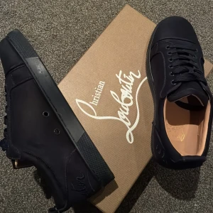 Svarta sneakers - Snygga svarta sneakers med klassisk röd sula och diskret broderad logga på hälen. Skorna har rund tå, snörning och är tillverkade i en mix av skinn och textil. Perfekta för dig som vill ha en stilren men lyxig look.