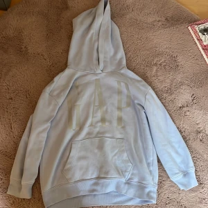 Ljusblå hoodie från GAP - Snygg ljusblå hoodie från GAP med stor logga framtill. Tröjan har huva, magficka och ribbade muddar vid ärmslut och nederkant. Perfekt för chill dagar och enkel att matcha med jeans eller joggers.