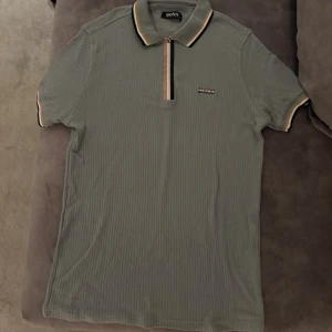 Ribbad pikétröja från Hugo Boss - Snygg ribbad pikétröja från Hugo Boss i mörk grönt med korta ärmar. Tröjan har dragkedja framtill, klassisk krage med beige och svart randig detalj samt diskret Boss-logga på bröstet. Perfekt för en clean och stilren look.