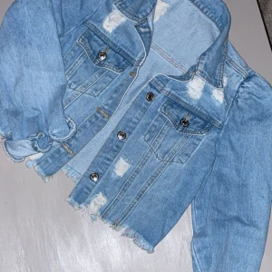 Croppad ljusblå jeansjacka med slitningar - Snygg croppad jeansjacka i ljusblå denim med slitna detaljer och fransad nederkant. Jackan har puffiga axlar, långa ärmar och silverfärgade knappar framtill. Två bröstfickor med lock och coolt distressed utseende som ger en trendig vibe. Knappast använd! 