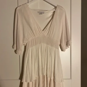 Ljus omlottklänning - Supersöt ljus kortklänning från New Yorker med v-ringning och omlottkänsla. Klänningen har korta ärmar och volangig nederdel. Perfekt för sommarens alla tillfällen. Luftigt tyg som ger en skön känsla.